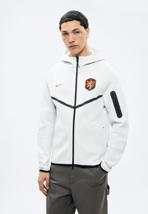 Noor mees, kes kannab valget lukuga kapuutsi, millel on lõvi embleem ja oranž Nike logo, sobitatud tumedate cargo-pükstega.