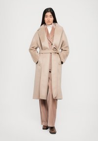 Light beige wool blend long coat with wide lapels, belt detailing, and side pockets, worn over a coordinating beige outfit and brown boots.
