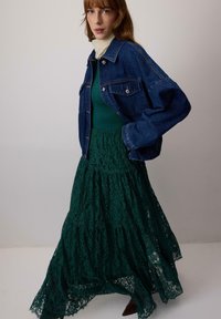 Giacca di denim sopra un abito maxi verde in pizzo. L'abito presenta strati a cascata e intricati motivi in pizzo. Lo sfondo è neutro.