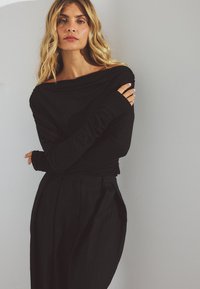 Zwarte lange mouw top met een off-shoulder ontwerp en aansluitende mouwen, gecombineerd met zwarte plooibroeken met hoge taille, gepresenteerd tegen een neutrale muur.