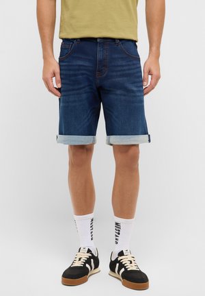 Mann trägt hochgekrempelte dunkelblaue Jeansshorts, weiße Socken mit schwarzem Schriftzug und schwarz-weiße Turnschuhe vor schlichtem Hintergrund.