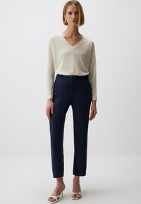 Weißes Langarm-V-Ausschnitt Oberteil kombiniert mit navyfarbener, taillierter Hose. Die Hose hat einen Knopfverschluss; das Modell trägt weiße Absätze. Glatte Texturen.