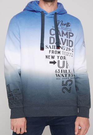 Man draagt een hoodie met een blauwe naar witte verloopkleur, zwarte tekst en nautisch geïnspireerde afbeeldingen, gecombineerd met donkerblauwe jeans.