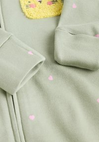 Zöld anyag, pink szívmintákkal, amelyen egy textúrázott sárga állatfej applikáció található a mellrészen, valamint hajtogatott mandzsetták a sleeves.