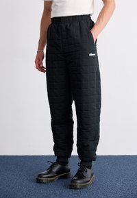 Pantalons matelassés noirs avec une taille élastique, dotés de poches latérales et d'un logo blanc. Portés avec des chaussures noires sur un tapis bleu.