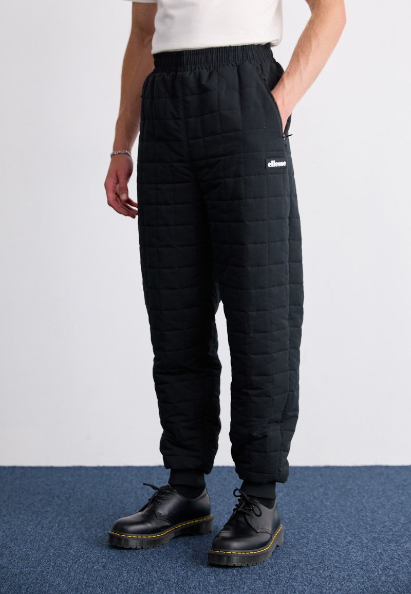 Pantalons matelassés noirs avec une taille élastique, dotés de poches latérales et d'un logo blanc. Portés avec des chaussures noires sur un tapis bleu.