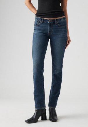 Kvinde iført mørkeblå slim-fit jeans, sort ærmeløs top og sorte ankelstøvler med hæl, stående på en lys baggrund.