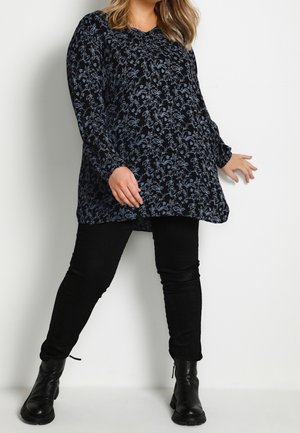 Vrouw die een zwarte bloemenprint blouse met lange mouwen draagt, zwarte broek en zwarte enkellaarzen, poserend tegen een witte muur.