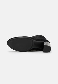 Botín de ante negro hasta el tobillo con punta redondeada suave, acento texturizado en el tacón y suela de goma plana. Logotipo presente en el tacón.