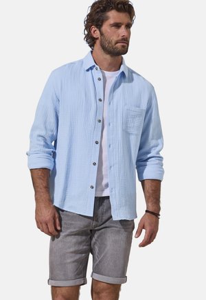 Man wearing light blue textured button-up shirt over white tee, gray rolled-up shorts, standing facing right on white background.