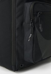 Sac à dos en tissu noir avec surface texturée, plusieurs poches zippées et accents en maille. Comprend des coutures renforcées et des zips durables.