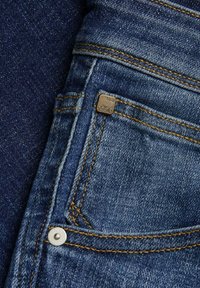 Jeans in denim con un lavaggio blu scuro, con cuciture dorate, un piccolo etichetta metallica con la scritta "JSN 1990" e una tasca con un bottone a pressione argentato.