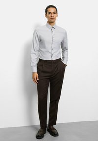 Gestreiftes hellgraues Hemd mit Button-Down-Kragen, kombiniert mit dunkelbraunen, plissierten Hosen und dunkelbraunen Slippern. Model steht vor einfarbigem Hintergrund.