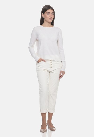 Donna in piedi con le mani nelle tasche, che indossa un maglione bianco a maniche lunghe, pantaloni bianchi corti con bottoni e tacchi beige su uno sfondo neutro.