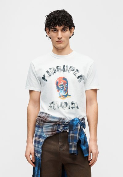 Λευκό βαμβακερό t-shirt με πολύχρωμο γραφικό τύπωμα και το κείμενο "KIDSUPER STUDIO!", συνδυασμένο με μπλε και καρό φανέλα φλάναλ που δένεται στη μέση.