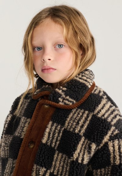Jeune personne aux cheveux clairs portant un manteau en laine à carreaux noir et beige avec des finitions en velours côtelé marron, regardant droit devant avec une expression neutre.