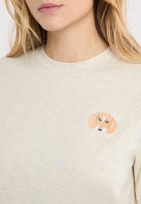 Hellbeiges Baumwoll-T-Shirt mit Rundhalsausschnitt, das auf der linken Brust ein besticktes Dackelkopf-Motiv in Braun, Beige und Schwarz zeigt.