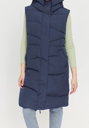 Frau trägt eine marineblaue, lange, gesteppte Steppweste mit Kapuze über einem hellgrünen Langarmshirt und hellblauen Jeans.