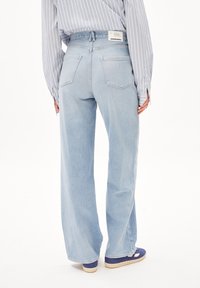 Person, der bærer lyseblå vide jeans, stribet skjorte stoppet ind og violet sneakers, står mod en hvid baggrund, set bagfra.