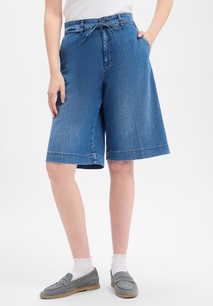 Vrouw die knielange blauwe denimshorts draagt, witte sokken, grijze loafers en een wit topje, staand met handen in en nabij de zakken.