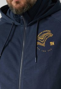 Marineblauwe hoodie met ritssluiting, gemaakt van zacht stof, met een hoge kraag, verstelbare trekkoorden en een gouden afbeelding op de borst.