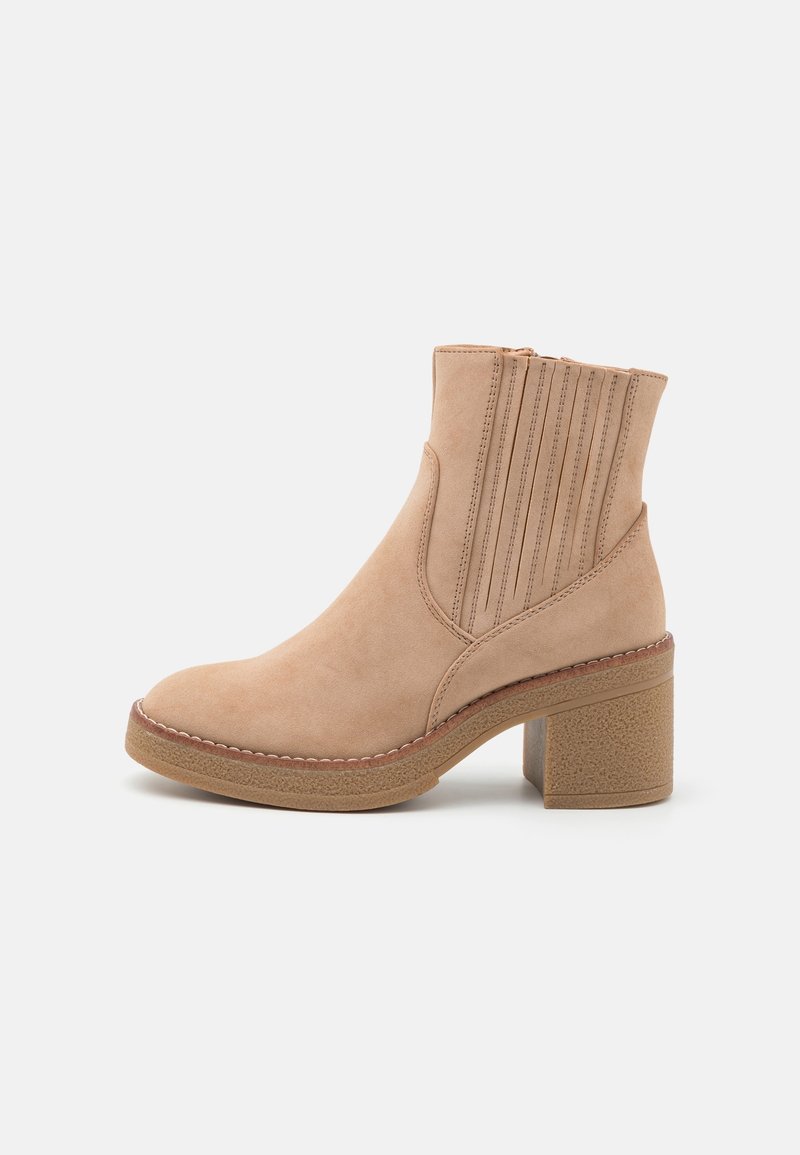Beige mockasins ankelboot med klack, dekorativ vertikal sömnad, rund tå och gummisula på vit bakgrund.