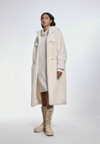 Kremowy, oversize'owy płaszcz z kapturem, o teksturowanym wykończeniu i przednimi kieszeniami, zestawiony z beżowymi botkami na sznurówki.
