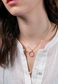 Gold necklace with a round pendant featuring floral accents and tiny colored stones, worn over a textured white blouse.