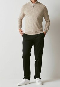 Beżowy sweter z zamkiem przy szyi o teksturowanym splocie, zestawiony z czarnymi spodniami i białymi trampkami. Strój cechuje się luźnym krojem i prostym designem.