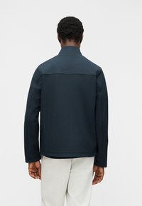 Άνδρας με την πλάτη γυρισμένη, φοράει σκούρο μπλε μπουφάν με ψηλό γιακά και ανοιχτό μπεζ παντελόνι, σε λευκό φόντο.