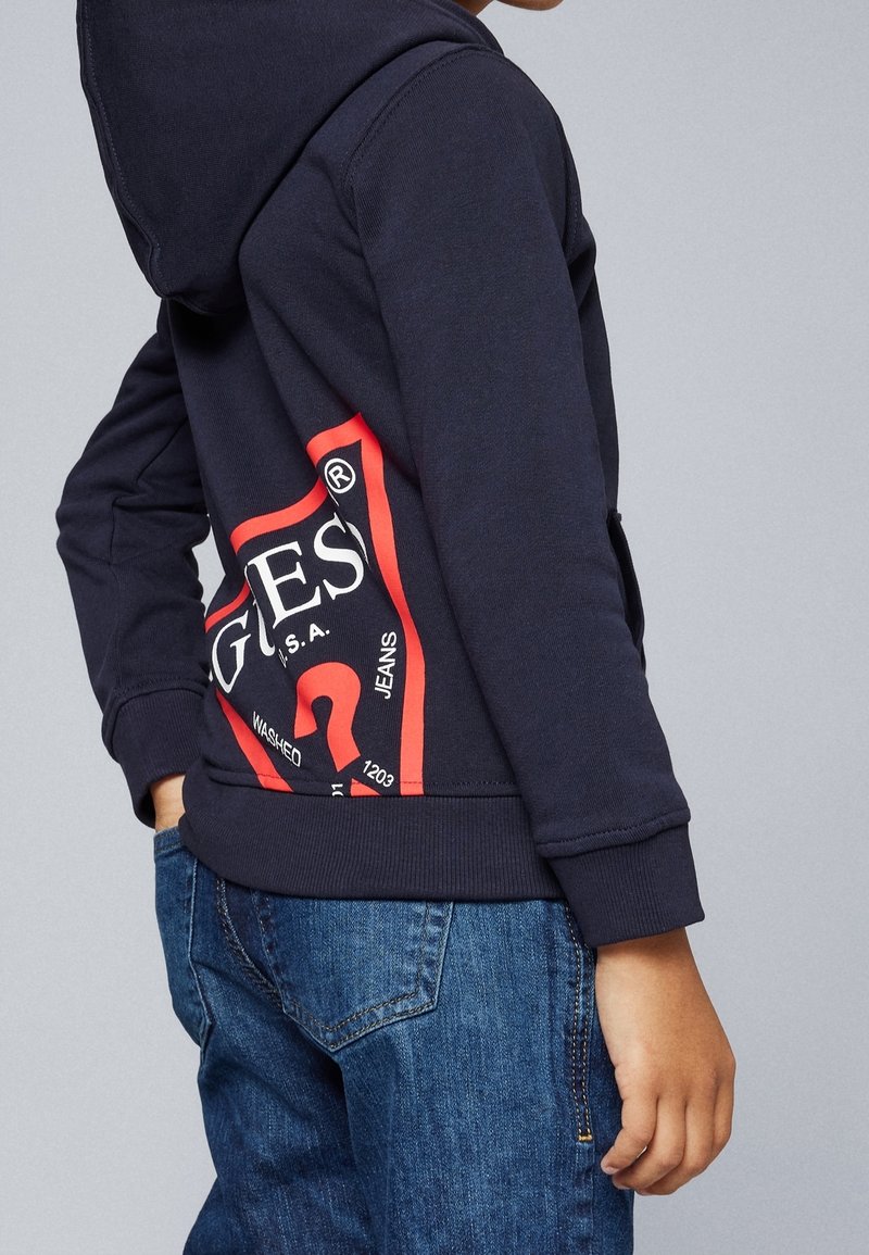 Enfant portant un sweat à capuche bleu marine foncé avec un logo Guess rouge et blanc sur le côté, associé à un jean en denim bleu, sur un fond uni.