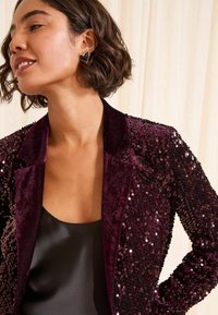 Burgundy sequined blazer with a smooth black top beneath. The jacket features a shawl collar and textured sequin details throughout.