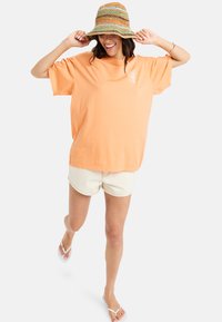 Oversize-Orange-Cotton-T-Shirt mit kurzen Ärmeln. Dazu beigefarbene Shorts und einen geflochtenen Hut mit mehrfarbigen Streifen. Flip-Flops sind inklusive.