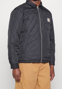 Černá prošívaná bunda se zipem vpředu, špičatým límcem, elastickými manžetami a náprsní kapsou s logem. Vyrobena z lehké látky.