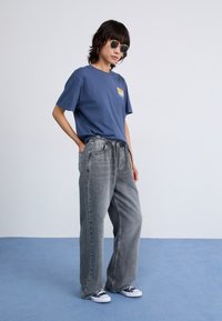Blå bomullst-skjorte med grafisk design, kombinert med løse grå denimbukser. Tilbehør inkluderer runde solbriller og blå sneakers.