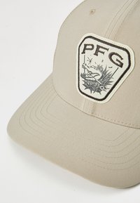 Căciulă din material bej, cu un vârful triunghiular având un design cu un crocodil alb-negru și textul "PFG." Margină curbată și detalii cusute vizibile.