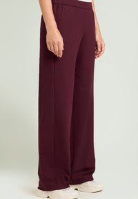 Pantalones de pierna ancha burdeos hechos de una tela suave y elástica; cuentan con una cintura elástica y bolsillos laterales, combinados con zapatillas blancas.