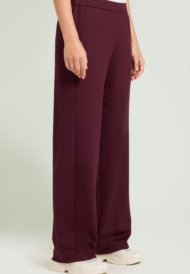 Pantalones de pierna ancha burdeos hechos de una tela suave y elástica; cuentan con una cintura elástica y bolsillos laterales, combinados con zapatillas blancas.