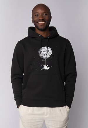 Lächelnder Mann trägt schwarzen Hoodie mit Grafik eines Astronauten, der im Weltraum schwebt und an einem großen Mondballon befestigt ist.
