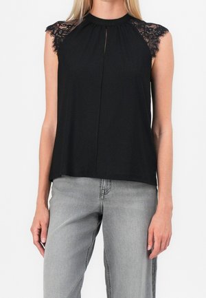 Kvinde iført en ærmeløs sort top med blonde-skuldre, parret med lysegrå jeans mod en hvid baggrund.