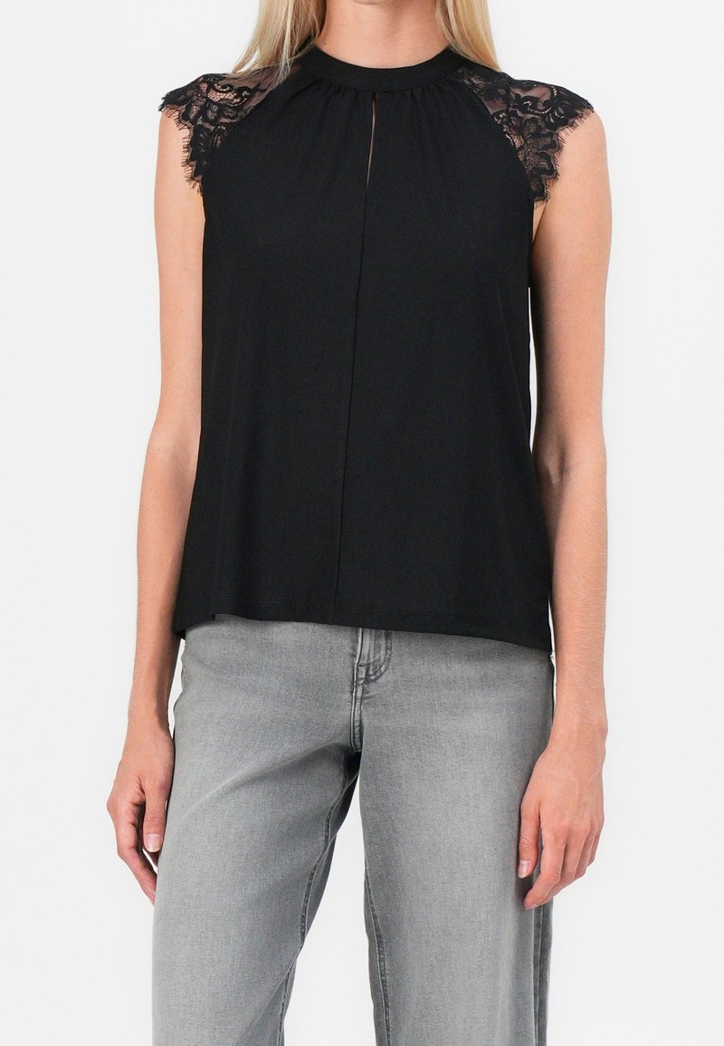 Donna che indossa un top senza maniche nero con spalline di pizzo, abbinato a jeans grigio chiaro, contro uno sfondo bianco.