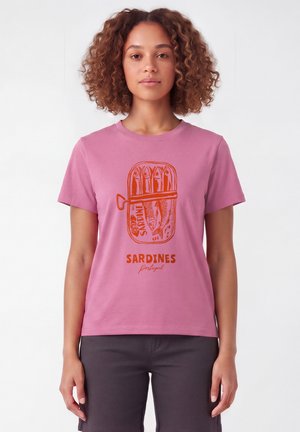 Frau mit lockigem Haar, die ein pinkes T-Shirt mit orangefarbener Sardinendosen-Grafik und Text "Sardines Portugal" trägt, vor weißem Hintergrund stehend.