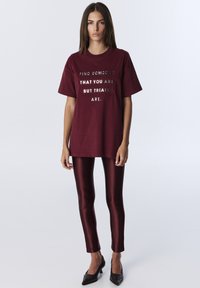 Bordowa koszulka z odblaskowym srebrnym napisem; zestawiona z błyszczącymi bordowymi legginsami i czarnymi szpiczastymi szpilkami. Gładki materiał, luźny krój.