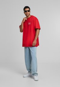 Rød oversized bomulds T-shirt med hvid tekst "Dead Wrong", parret med lyseblå denimbukser og hvide sneakers. Modellen bærer sorte solbriller.