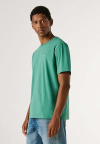 Jeune homme portant un t-shirt vert à manches courtes et un jean bleu, debout avec une expression neutre sur un fond clair uni.