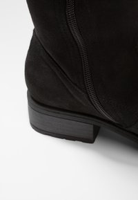 Primer plano del tacón de una bota de ante negra con costuras visibles y suela de goma sobre un fondo blanco.