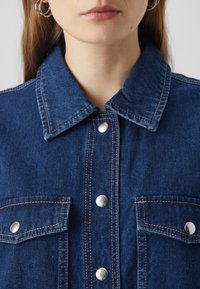 Veste en denim bleu foncé avec un col, des poches avant, des boutons-pression en argent et des détails de surpiqûre visibles. Texture de tissu lisse.