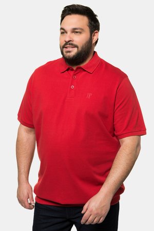 Bearded man wearing a red short-sleeve polo shirt and dark pants, standing against a plain light background, looking slightly right.