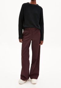 Osoba oblečená v černém topu s dlouhým rukávem, tmavě vínových širokých kalhotách a bílých platformových teniskách stojící před jednoduchým pozadím.