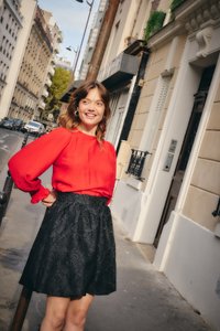 Junge Frau in roter Bluse und schwarzem Blumrock steht lächelnd auf dem Bürgersteig einer Stadt mit europäisch geprägten Gebäuden und parkenden Autos in der Nähe.